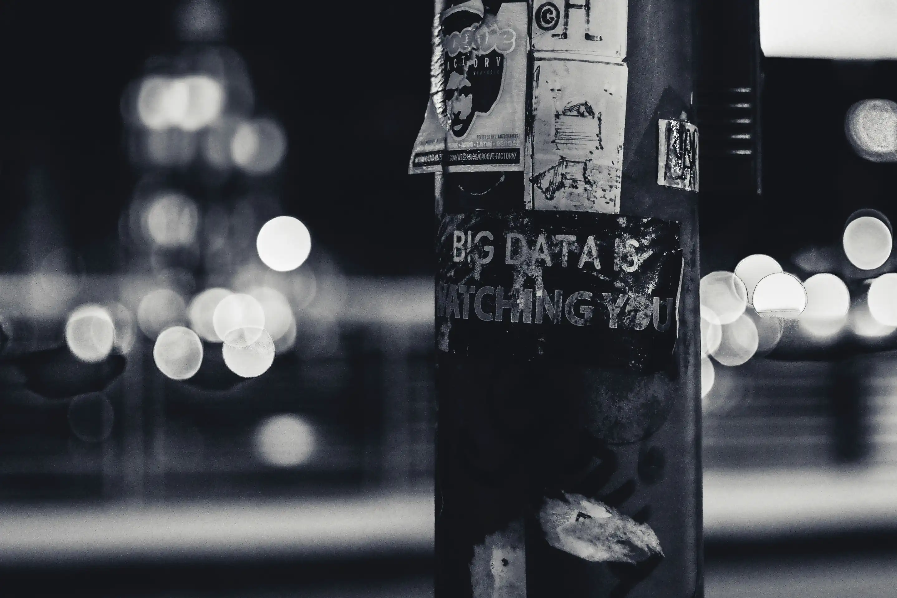 Black and white photo of a street post at night. The street post has some ripped stickers on it and a stencilled graffiti saying Big Data is Watching You.